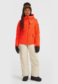 Felle oranje geïsoleerde jas met een capuchon, ritssluiting aan de voorkant en twee zakken, gecombineerd met crèmekleurige broek en zwarte handschoenen en laarzen.