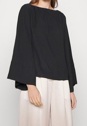 Femme portant un chemisier noir ample à manches longues avec de larges poignets et un pantalon plissé taille haute beige clair, debout devant un fond uni.