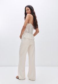 Čipkový peplum top v smotanovej farbe s tenkými ramienkami, kombinovaný s širokými béžovými nohavicami. Hladká textúra s viacvrstvovým detailom na leme.