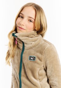 Beige fleece jack met hoge kraag, teal rits en geborduurde patch met bergen. Zachte textuur en een rits aan de voorkant.