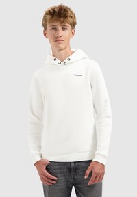Tienerjongen in een witte hoodie met de tekst "BALLIN", staand met zijn handen dicht bij zijn zakken tegen een effen lichte achtergrond.