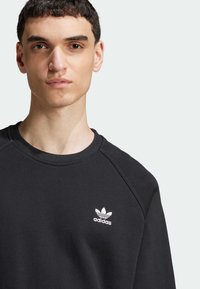 Felpa nera con scollo rotondo e maniche raglan. Presenta un piccolo logo Adidas bianco sul petto, realizzata in tessuto testurizzato.