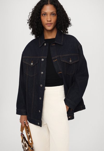 Donkerdenim jack met zilveren knopen, twee borstzakken en oranje stiksel, gecombineerd met een zwarte top en lichtgekleurde broek.