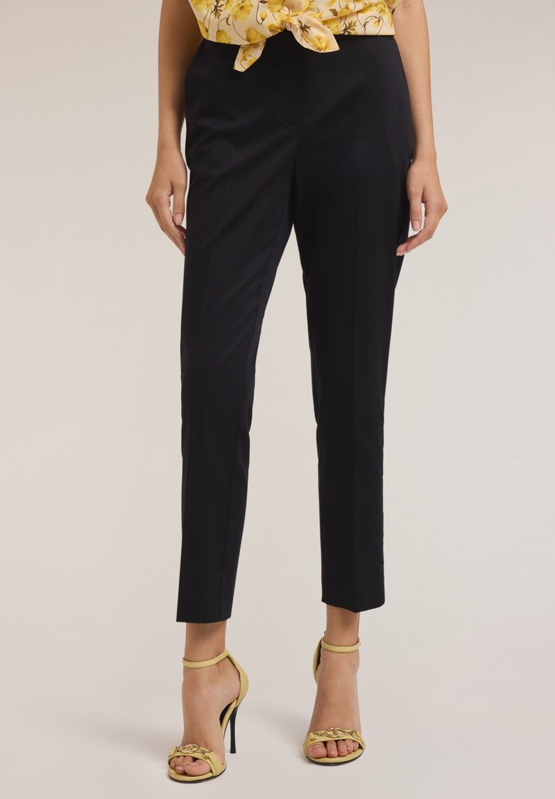 Pantalons noirs sur mesure à coupe ajustée, présentant une texture lisse et des coutures minimales. Portés avec une chemise à fleurs jaunes et des talons jaunes.