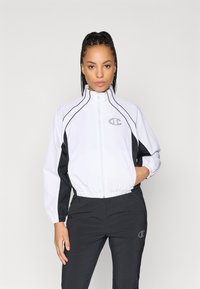 Balta un melna vēja necaurlaidīga jaka ar augstu apkakli, ar rāvējslēdzēju un elastīgām aprocēm, ar logo uz krūtīm un sānu akcentu svītrām.