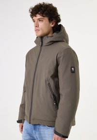 Chaqueta aislante de color verde oliva con cremallera frontal, cuello alto, capucha ajustable y bolsillos laterales con cremallera. Presenta un logotipo de la marca en la manga.