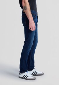 Mann trägt dunkelblaue Slim-Fit-Jeans, weiße Sneakers mit schwarzen Streifen und ein schwarzes T-Shirt, steht seitlich vor einem einfarbigen Hintergrund.