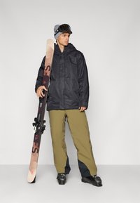 Veste noire imperméable avec capuche, pantalon kaki, lunettes de ski, tenant des skis avec fixations. Détails notables incluent le contraste de couleurs et les textures.