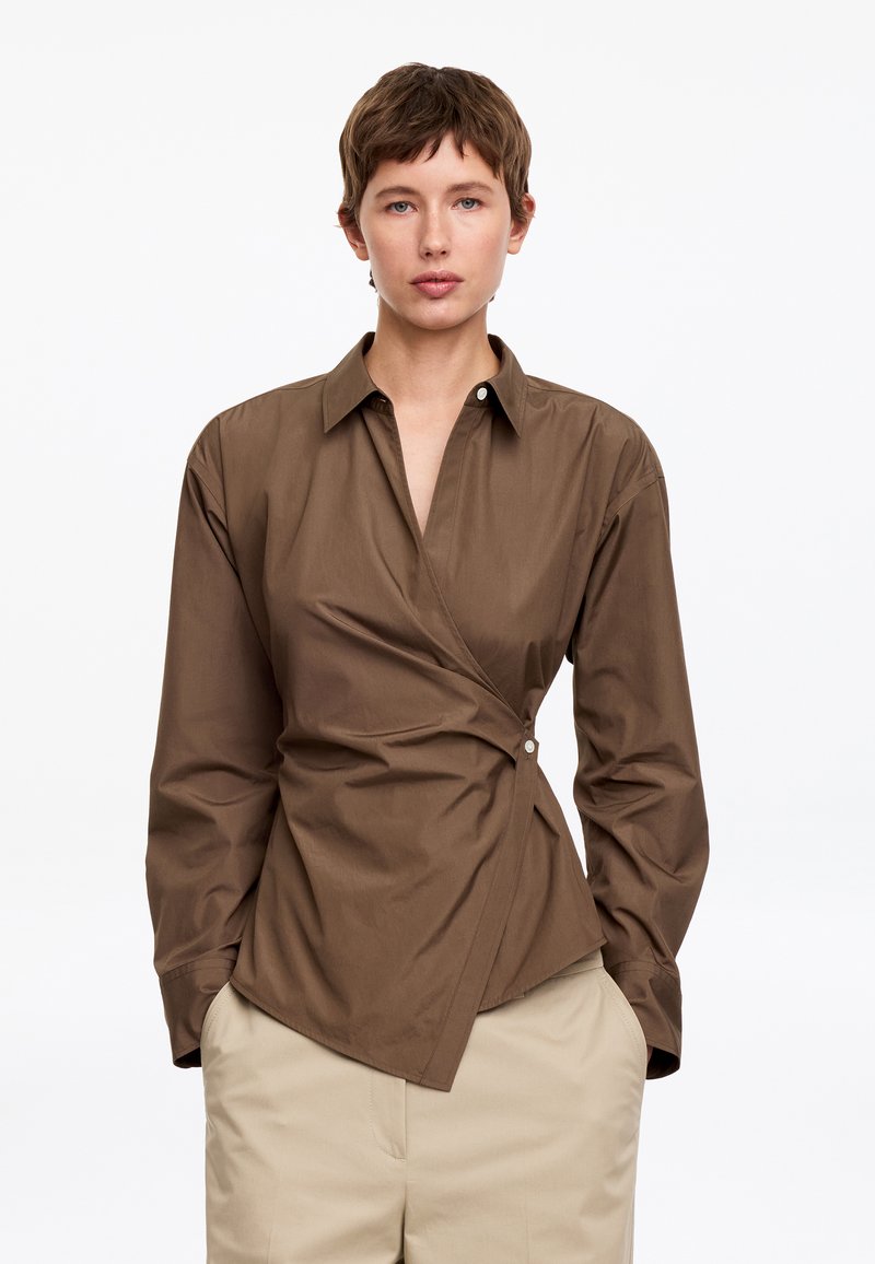 Personne aux cheveux courts et bruns portant une chemise marron style cache-cœur et un pantalon beige, debout les mains dans les poches sur fond blanc.