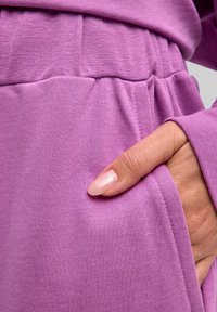 Fioletowy bawełniany element garderoby o luźnym kroju, z teksturowanym elastycznym pasem i boczną kieszenią, trzymany w ręce z zadbanymi, pomalowanymi paznokciami.