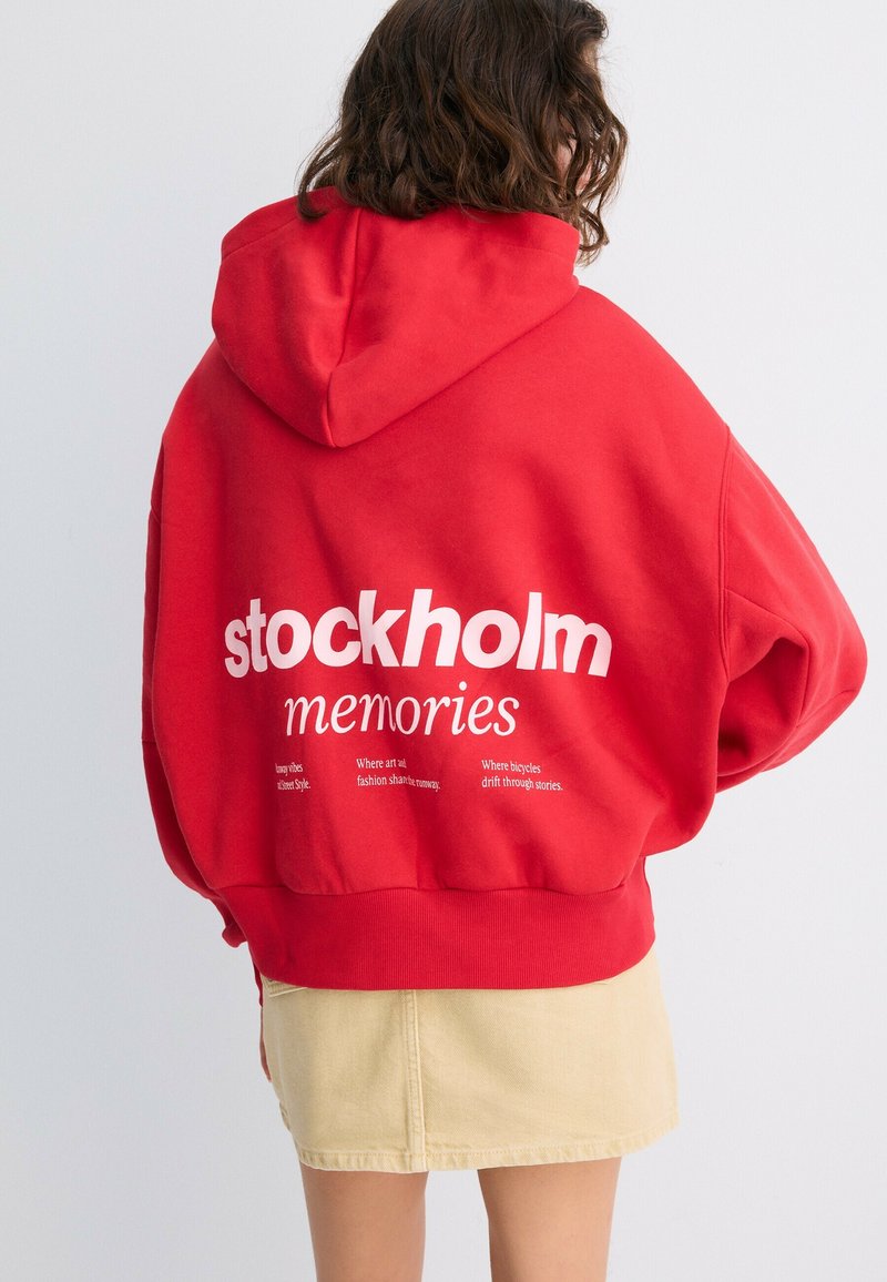 Personne portant un sweat à capuche rouge avec la capuche baissée et un texte blanc "stockholm memories" dans le dos, associé à une jupe beige, tournée dos à la caméra.