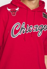 Sudadera roja con capucha que presenta "Chicago" en una escritura blanca, con un logo de los Bulls en negro y rojo en el hombro izquierdo. Cuerdas ajustables y una textura suave.