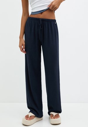 Frau trägt locker sitzende marineblaue Kordelzughose und weiße Plateausandalen, hebt ein weißes, geripptes Tanktop hoch, um die Taille zu zeigen.