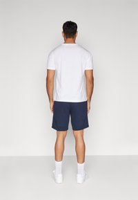 Camiseta de algodón blanca y pantalones cortos azul marino, con cintura elástica. Combinados con calcetines blancos y zapatillas, vistos desde atrás.