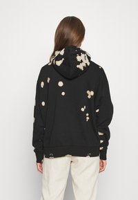 Czarna bluza z kapturem z plamami po wybielaczu, wyposażona w kaptur ze sznurkiem, długie rękawy oraz ściągacz na dole. Wykonana z miękkiego materiału dla komfortu.