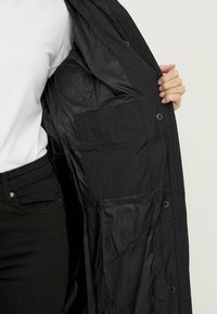 Zwarte gewatteerde jas met een gladde nylon binnenkant, voorzien van een grote binnenzak en knoopsluitingen aan de voorkant voor het sluitmechanisme.