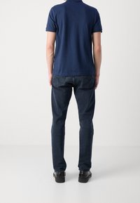 Námořnická modrá polo košile s krátkým rukávem ve spojení s tmavě modrými džíny. Džíny mají vyšitá zadní kapsy a černé boty.