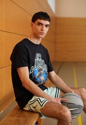 T-shirt grafico nero con design blu e bianco, abbinato a pantaloni corti da basket verde menta con accenti neri. Tiene una palla da basket grigia.