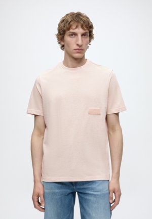Jeune homme aux cheveux blonds ondulés portant un t-shirt rose clair à manches courtes et un jean bleu sur un fond blanc uni.