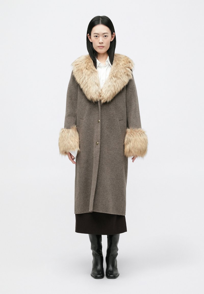 Καφέ μάλλινο παλτό με μεγάλο κολάρο και μανίκια από ψεύτ�ικη γούνα. Διαθέτει ευθεία γραμμή, δύο μπροστινές τσέπες και μεταλλικά κουμπιά.