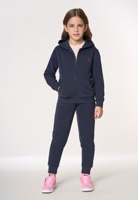 Felpa con zip blu navy e pantaloni da jogging abbinati in tessuto morbido con due tasche frontali. Presentano un piccolo logo rosso e sneakers rosa con suole bianche.