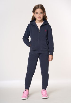 Felpa con zip blu navy e pantaloni da jogging abbinati in tessuto morbido con due tasche frontali. Presentano un piccolo logo rosso e sneakers rosa con suole bianche.