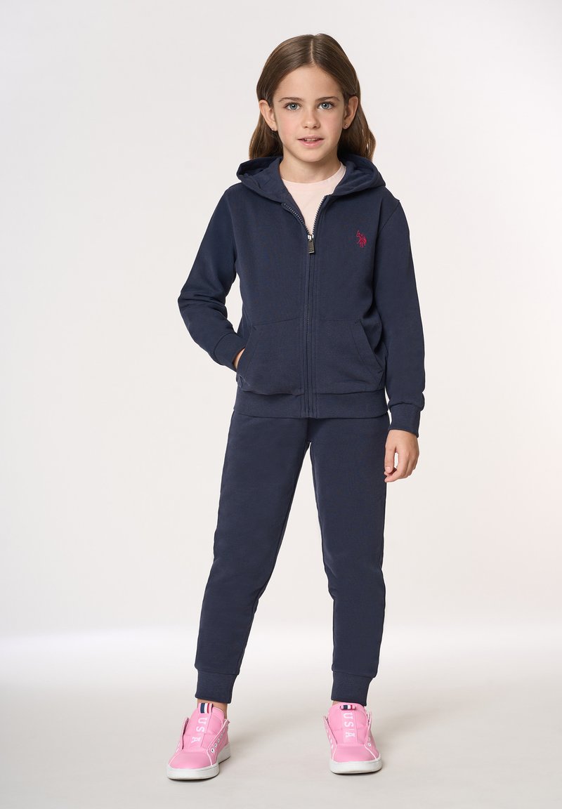 Felpa con zip blu navy e pantaloni da jogging abbinati in tessuto morbido con due tasche frontali. Presentano un piccolo logo rosso e sneakers rosa con suole bianche.