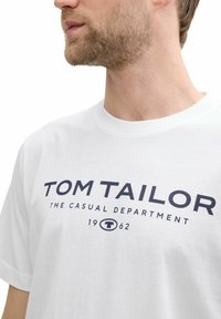 Weißes Baumwoll-T-Shirt mit kurzen Ärmeln, das auf der Vorderseite mit einem navyblauen Druckgrafik versehen ist, auf dem "TOM TAILOR THE CASUAL DEPARTMENT 1962" steht.