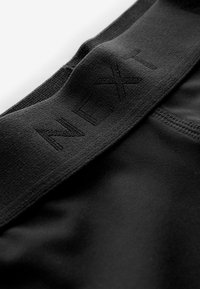 Zwarte elastische tailleband met een geperst "NEXT" logo, bevestigd aan een gladde zwarte stof. Naaddetails zichtbaar aan de rand van het kledingstuk.