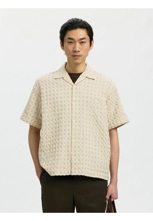 Junger Mann mit dunklen Haaren trägt ein beiges, strukturiertes kurzärmeliges Hemd über einem braunen Top und hält eine braune Tasche mit einer Hand.