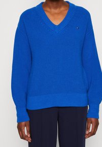 Pull en maille bleu avec col en V, manches larges et poignets côtelés. Présente un motif texturé et un petit logo sur la poitrine.