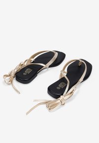 Las sandalias planas doradas cuentan con un diseño minimalista, suela negra, detalle de tiras cruzadas y acentos de cinta atada en la parte trasera.