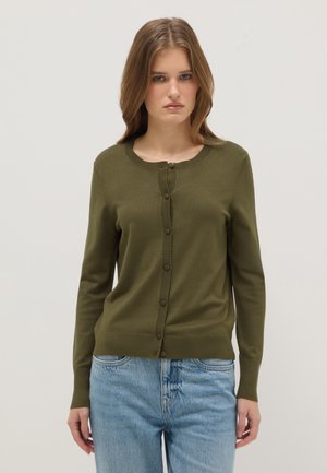 Mujer con cárdigan abotonado de color verde oliva y jeans azul claro, de pie frente a un fondo claro y liso.