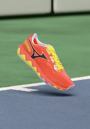 Chaussure de sport orange vif avec des lacets jaunes et des accents de semelle, logo noir sur le côté, flottant au-dessus d'un terrain de sport bleu et vert.