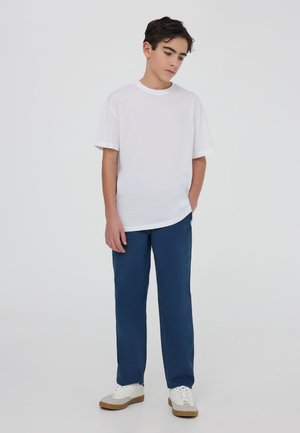 Biała koszulka z krótkim rękawem o luźnym kroju, zestawiona z granatowymi spodniami o prostych nogawkach oraz białymi trampkami z beżowymi podeszwami, na neutralnym tle.