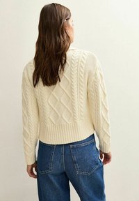 Crème kabelgebreide trui met een cropped fit, voorzien van een geribbelde zoom en verschillende textuurpatronen. G gedragen met blauwe high-waisted jeans.