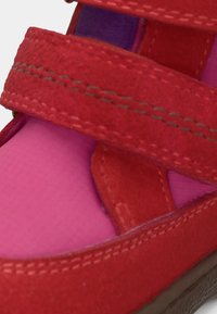 Närbild av röd mockaskosnör med synlig söm över rosa tyg och gummiundersidans kant.