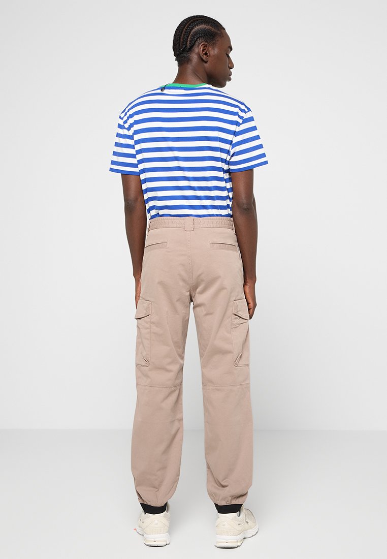 Man staat met de rug naar de kijker gekeerd en draagt een blauw met wit gestreept kortarmshirt, beige cargobroek en witte sneakers.