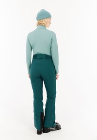 Teal langesleeve top met hoge kraag en groene skibroek met een zwart paneel bij de enkel, gecombineerd met zwarte skiboots.
