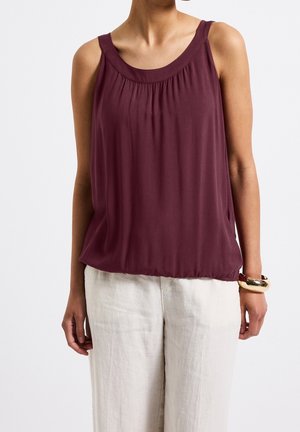 Kobieta w bezrękawnym bordowym topie z marszczonym dekoltem i luźnych białych spodniach, uzupełniona złotą bransoletką na prawej ręce.