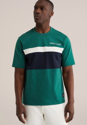 Mees, kes kannab roheline-valge-sinimustvalge värvikombinatsiooniga T-särki, millel on tekst „Leisure Court Club", valgete pükstega, seisab neutraalse tausta ees.