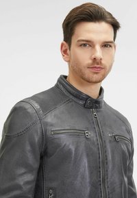 Giacca di pelle nera con colletto alto, dettagli a coste sulle spalle e due tasche sul petto con zip. Texture liscia con un leggero lucido.