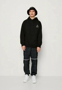 Young person standing wearing a black hoodie with a white triangle logo, plaid pants with white stripes, white sneakers, and a black bucket hat.