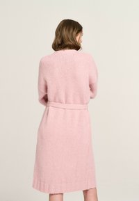 Rosa gestrickte Strickjacke mit Gürtel, die eine weiche Textur, lange Ärmel und einen mittellangen Saum aufweist, von hinten betrachtet.