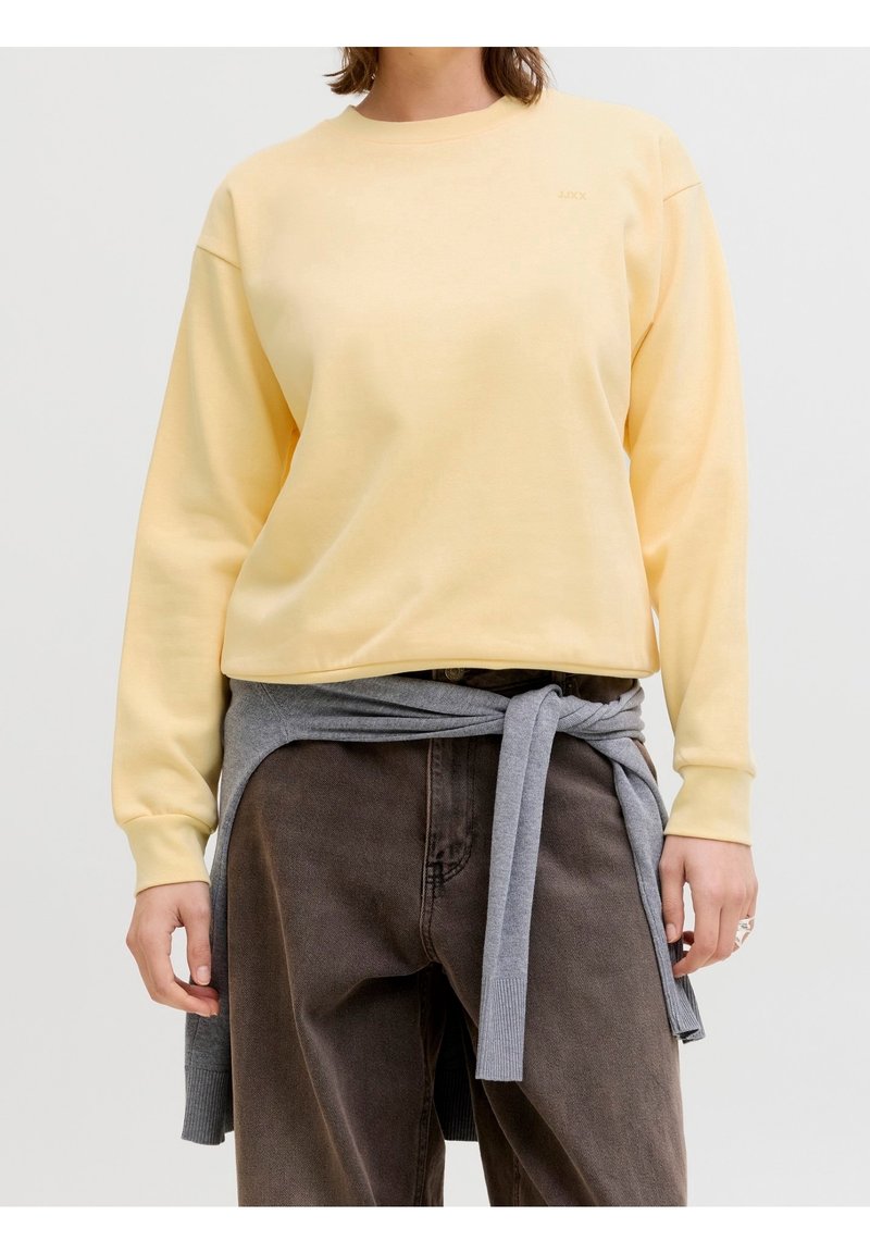 Personne portant un haut à manches longues jaune pâle et un pantalon brun foncé avec un pull gris noué autour de la taille, debout devant un fond neutre.