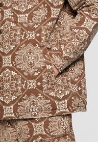 Brunt mønstret stof med intrikate hvide design, herunder blomster- og geometriske motiver. Glat tekstur med et lagdelt udseende.