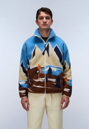 Hombre que lleva una chaqueta de forro polar azul con dibujos de montañas, árboles y cabañas, combinada con pantalones de color crema, posando frente a un fondo neutro.