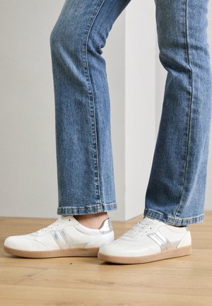 Białe skórzane sneakersy ze srebrnymi akcentami noszone z niebieskimi dżinsami, stojące na drewnianej podłodze wewnątrz pomieszczenia.