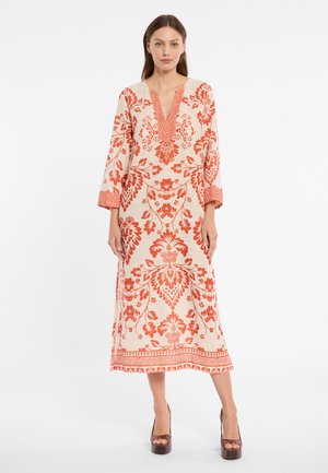 Frau trägt knöchellanges cremefarbenes Kleid mit orangefarbenen Blumenmustern und dreiviertel langen Ärmeln, stehend vor schlichtem weißem Hintergrund.