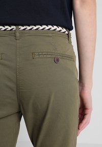 Olivgröna byxor med en slät textur, som har en bakficka med en brun knapp. Ett vävt bälte i marinblått och krämfärgat betonar midjan.